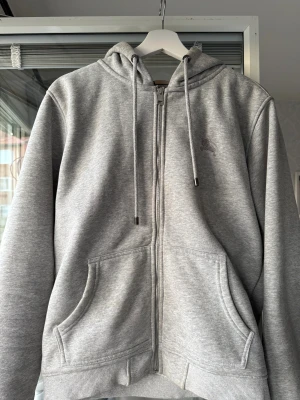 Grå hoodie från Burberry med dragkedja - Säljer en grå hoodie från Burberry i storlek M med klassiskt rutigt foder i huvan. Tröjan har hel dragkedja, snörning vid huvan och två stora fickor framtill. Materialet är mjukt med inslag av bomull och polyester, perfekt för chill dagar.