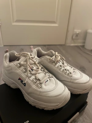 Vita chunky sneakers från Fila - Lite använda men i got skick! Säljer ett par vita chunky sneakers från Fila med platt sula och snörning. Skorna har rund tå och är tillverkade i syntetmaterial med Fila-logga i blått och rött på sidan och tungan. Perfekt för dig som gillar sportig och trendig stil. 