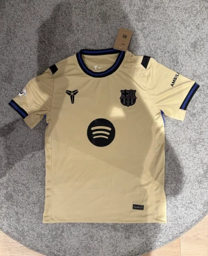FC Barcelona Raphinha 11 bortatröja - Snygg beige FC Barcelona bortatröja från Nike med Raphinha 11 på ryggen. Tröjan har svarta och blå detaljer, klubbmärke och sponsorloggor på bröstet samt tryck på ärmarna. Tillverkad i lätt polyester som andas, perfekt för fotboll eller att bära casual.