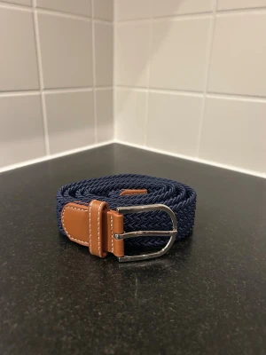 Marinblå flätad bälte med läderdetaljer - Snyggt marinblått flätat bälte med bruna läderdetaljer och silvrigt spänne. Bältet har en stilren design med vit söm på lädret och passar perfekt till jeans eller chinos. Materialet är en kombination av textil och läder för både komfort och stil. 105 cm men töjbart och passar bredare 