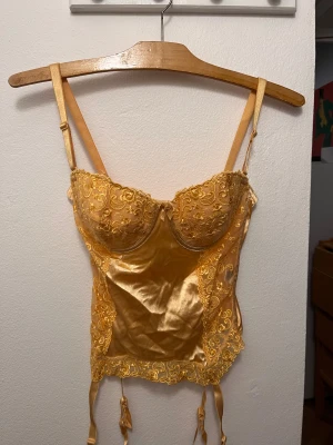 Gul korsett med spets och satin - Snygg gul korsett med justerbara axelband och dekorativa spetsdetaljer framtill. Materialet är glansig satin med broderad spets över kuporna och längs kanterna. Korsetten har hyskknäppning bak och fästen för strumpeband. 