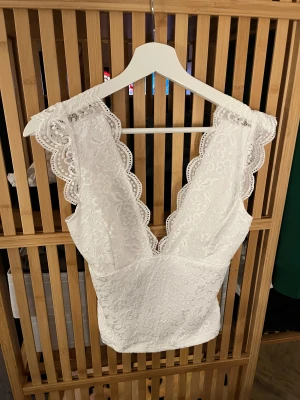 Vit spetsblus med djup v-ringning från SHEIN - Supersöt vit blus från SHEIN i storlek S, med djup v-ringning och breda axelband. Blusen är täckt av blommig spets och har fina vågiga kanter. Den är figurnära och har öppen rygg med liten knytning bak. Perfekt för dig som gillar romantisk och feminin stil.