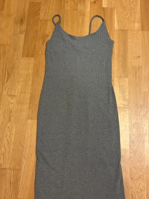 Grå ribbad långklänning med smala axelband - En stilren grå långklänning med smala axelband och ribbad struktur. Klänningen har en kroppsnära passform och är ärmlös, vilket ger en enkel och modern look. Perfekt för dig som gillar minimalistisk stil.