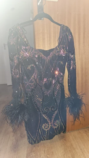 Svart glittrig paljettklänning med fjädrar från Fashion Nova - Unik svart kortklänning med glittriga paljetter i lila och guld, långärmad med dramatiska fjäderdetaljer vid ärmsluten. Klänningen har ett mönstrat broderi och markerad midja, perfekt för dig som vill sticka ut.