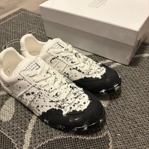Maison Margiela svartvita sneakers - Unika sneakers från Maison Margiela i vitt tyg med svart färgstänksmönster. Skorna har snörning, rund tå och platt sula. Kommer med originalkartong och vita skyddande överdrag. Perfekt för dig som gillar statement pieces och cool design. Öppen för byten