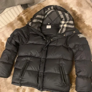 Svart pufferjacka från Burberry - Säljer en svart pufferjacka från Burberry med klassisk rutig huva i grått och vitt. Jackan har dragkedja och knappar framtill, två sidofickor med dragkedja och en snygg Burberry-logga på ärmen. Perfekt för kalla dagar och riktigt stilren.