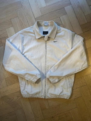 Vit manchester vindjacka från Nike - Säljer en stilren vit vindjacka från Nike i mjuk manchester. Jackan har klassisk krage, svart broderad swoosh på bröstet och dragkedja framtill. Elastiska muddar vid ärmslut och nederkant ger en skön passform. Pris är inte fast så bjud gärna. Det passar ganska stor i storlek så passar jättebra i S