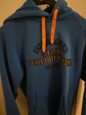 Blå hoodie Peak Performance S - Blå hoodie från Peak Performance i storlek S med orange snören och broderad logga på bröstet. Tröjan har huva, magficka och coolt tryck med Established 1986. Perfekt för chill eller sportiga dagar.