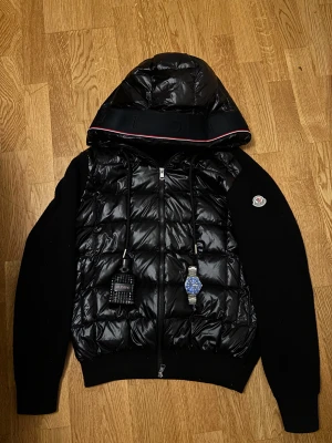 Svart Moncler pufferjacka med huva - Svart pufferjacka från Moncler med quiltad front och stickade ärmar. Jackan har en stor huva med snygg kantdetalj, dragkedja framtill och Moncler-logga på ärmen. Materialmix av dun och stickad textil gör den både varm och stilren.