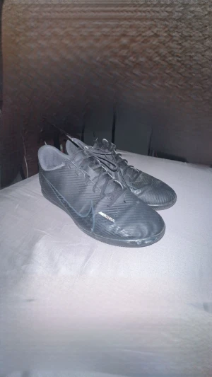 Svarta Nike Mercurial fotbollsskor - Säljer ett par svarta Nike Mercurial fotbollsskor med snyggt randigt mönster och stora Nike-loggor på sidorna. Skorna har snörning och platt sula, tillverkade i syntetmaterial för bra grepp och komfort på planen. Perfekt för dig som vill ha stil och funktion på träningen.