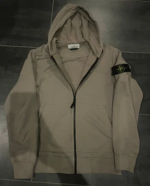 Beige hoodie från Stone Island - Säljer en beige hoodie från Stone Island med dragkedja och klassisk logga på vänster ärm. Tröjan har huva, känguruficka och är tillverkad i mjuk bomull. Perfekt för dig som gillar streetwear och vill ha något stilrent men ändå fett.