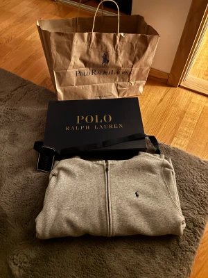 Grå zip hoodie från Polo Ralph Lauren - Snygg grå hoodie från Polo Ralph Lauren med dragkedja framtill och klassisk logga broderad på bröstet. Tröjan har huva och är långärmad, perfekt för chill dagar. Kommer med originalförpackning och tagg kvar.