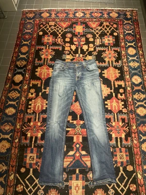 Blå nudie jeans straight fit - Säljer ett par snygga nudie jeans i klassisk blå tvätt med slitningar och lätt ruggad look. Modellen har raka ben, fem fickor och detaljerade sömmar på bakfickorna. Perfekt passform med normal midja och dragkedja i gylfen.