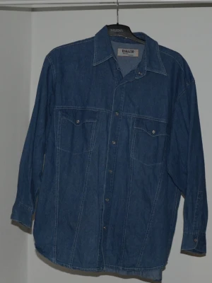 Blå jeansskjorta från Ragazzo - Säljer en klassisk blå jeansskjorta från Ragazzo med lång ärm och snygga knappar framtill. Skjortan har två bröstfickor med lock och en loose passform. Stl. L/XL