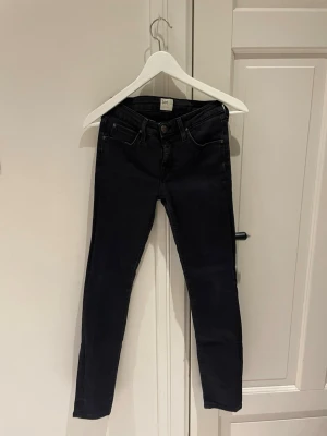 Svarta skinny jeans från Lee - Säljer ett par svarta skinny jeans från Lee med klassisk femficksdesign och metallknapp med logga. Jeansen har låg till normal midja och är tillverkade i stretchigt denim för en tight passform. Perfekta till sneakers eller boots.