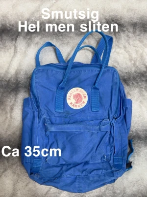 Blå Kånken ryggsäck från Fjällräven - Klassisk blå Kånken ryggsäck från Fjällräven, ca 35 cm hög. Ikonisk rund logga framtill, stort huvudfack med dragkedja och en mindre ficka på framsidan. Justerbara axelband och bärhandtag upptill. Perfekt för skola eller utflykter.🌸 Hel men sliten. Ca 35cm hög. Ej tvättad så smutsig.