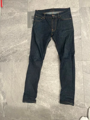 Nudie Jeans Lean Dean Dry  - Säljer ett par mörkblå Lean Dean jeans från Nudie Jeans i modellen Dry 16 Dips. De har klassisk femficksdesign, kontrastsömmar och är tillverkade i 99% bomull och 1% elastan för lite stretch. Snygg skinny passform och ikoniska Nudie-broderier på bakfickorna. Så gott som nya.Midja 42cm. Längd 105.