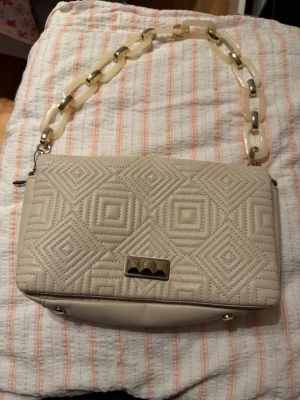 Beige quiltad axelväska med kedja - Säljer en beige axelväska med quiltat geometriskt mönster och snygg kedja i plast och metall. Väskan har en rektangulär form och ett coolt metallspänne framtill. Perfekt för dig som gillar trendiga detaljer och stilren design.