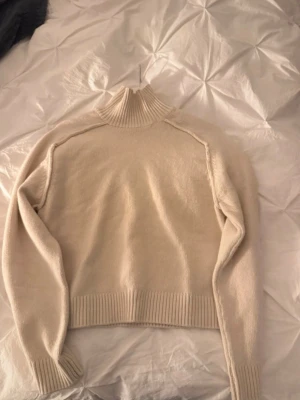 Beige polotröja i stickat material - Säljer en beige polotröja i stickat material med ribbade muddar och hög krage. Tröjan har en loose passform och är perfekt för kyliga dagar. Enkel och stilren design som är lätt att matcha med olika outfits.