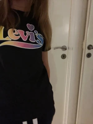Svart Levi's t-shirt med färgglatt tryck - Pris kan diskuteras!🩷Den ser lite fläckig ut men det bara bara för att jag tog bild med blixt!💕Svart t-shirt från Levi's med ett stort, färgglatt logotryck på bröstet i gul, rosa, lila och blå toner. Klassisk rund hals och korta ärmar. Materialet är mjuk bomull som känns skönt mot huden. Perfekt för dig som gillar streetstyle och vill ha en ikonisk look.