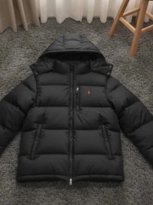 Svart pufferjacka från  Ralph  - Svart pufferjacka från  Ralph  med huva och två sidofickor med dragkedja. Jackan har en röd Polo-logga på bröstet och quiltad design. Perfekt för kalla dagar och har en snygg, sportig look.