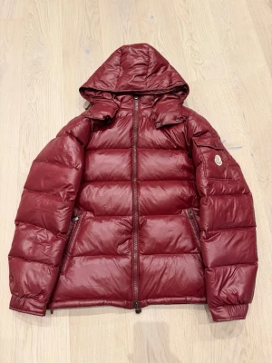 Vinröd Moncler dunjacka med huva - Säljer en vinröd dunjacka från Moncler med huva och dragkedja framtill. Jackan har puffad design, ficka med logotyp på ärmen och är tillverkad i polyester. Perfekt för kalla dagar och har en riktigt snygg look som sticker ut.