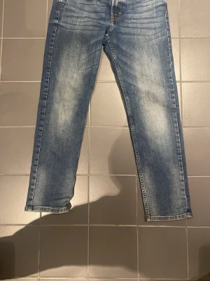 Blå tapered jeans från &Denim - Säljer ett par blå jeans från &Denim med relaxed tapered fit. Jeansen har klassisk femficksdesign, snygg tvätt med slitningar och är tillverkade i en mix av bomull och polyester. Perfekta för en chill och avslappnad look.