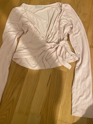 Ljusrosa omlottopp från Gina Tricot - Säljer en ljusrosa omlottopp från Gina Tricot i mjuk bomull. Toppen har långa ärmar och knyt i midjan för en snygg passform. Perfekt att styla med jeans eller kjol för en fräsch look. Fint skick, ej använd mycket. 