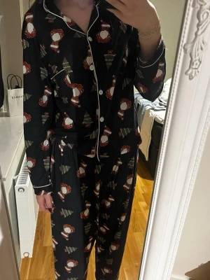 Svart julpyjamas sett med tomtemönster - Mysig svart pyjamas settmed knappar och vit piping, täckt av gulliga tomtar och julgranar. Långärmad och med klassisk krage, perfekt för att chilla under vinterns lediga dagar. 
