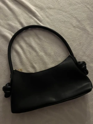 Svart axelväska med knutdetaljer - Säljer en stilren svart axelväska i skinnimitation med snygga knutdetaljer på axelremmen. Väskan har en mjukt rundad form och stängs med en guldfärgad dragkedja. Perfekt storlek för det viktigaste och passar till många olika looks. Onvänd,ny❗️