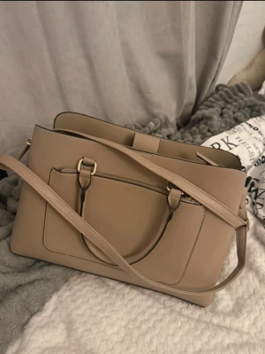 Beige väska från Primark - Snygg beige axelväska från Primark med stilren design och praktiska fack. Väskan har både handtag och justerbar axelrem, samt detaljer i guld. Materialet är slätt syntet och insidan har flera fack med dragkedja för smart organisering.