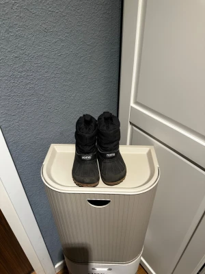 Barn vinterstövlar från Reima svarta  - Säljer ett par svarta vinterstövlar från Reima med grov brun sula och reflekterande detaljer baktill. Stövlarna har ett quiltat skaft i syntetmaterial och är perfekta för kalla dagar. Sulan har ett mönster som ger bra grepp på snö och is. Vattenavisande 