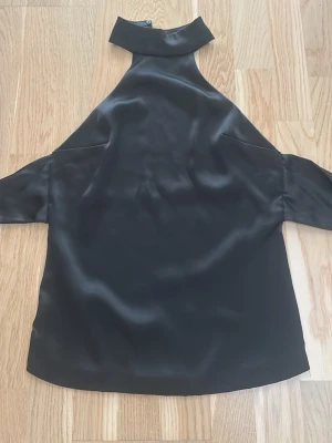 Svart omlott polotopp från Zara - Säljer en svart polotopp från Zara i slät satinliknande polyester. Toppen har omlott-design med stor rosett bak och knäppning i halsen. Ärmlös modell med hög krage som ger en cool och elegant vibe. Perfekt för dig som gillar stilrena detaljer. Mitenknapen är borta men fingerar ändå