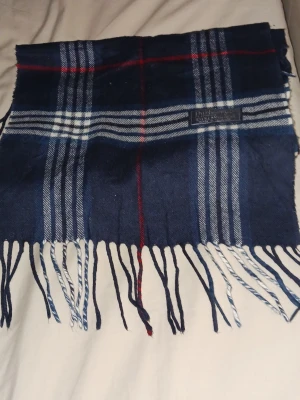 Burberrys scarf - Säljer en klassisk blårutig  Burberrys scarf, tillverkad i 100% lammull. Scarfen har ett snyggt rutmönster i blått, vitt och inslag av rött, samt fransar nedtill som ger en cool touch. 