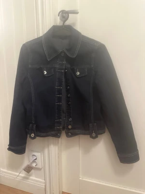 Mörkblå jeansjacka från Sara, stl 36 - Snygg mörkblå jeansjacka från Sara i storlek 36. Klassisk modell med krage, framfickor med knappar och kontrastsömmar. Jackan har långa ärmar och knäppning framtill med silverfärgade knappar. Perfekt för dig som gillar en clean och tidlös look.