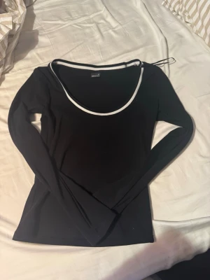 Svart långärmad topp från Gina Tricot - Svart långärmad topp från Gina Tricot med vit kant runt den djupa, rundade halsringningen. Topp i stretchigt material som sitter snyggt och passar perfekt till jeans eller kjol.
