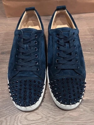Christian Louboutin skor - Säljer ett par mörkblå sneakers från Christian Louboutin i mocka med vita sulor och ikoniska röda undersidor. Skorna har snörning och är dekorerade med svarta nitar på tån för en edgy vibe. Perfekta för dig som vill sticka ut med lyxiga detaljer.