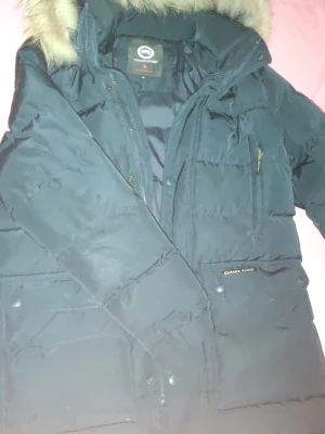 Canada goose M - Svart dunväst från Canada Goose med klassisk logga på bröstet och huva med pälsdetalj. Västen har dragkedja och tryckknappar framtill, samt flera praktiska fickor. Perfekt för kalla dagar och har en stilren, quiltad design. Och är använt i två veckor.