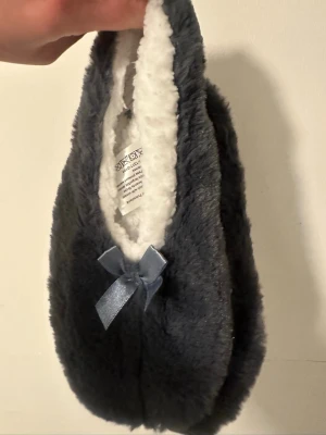 Grå fluffiga tofflor med rosett - Mysiga grå ballerinaskor från Rusta, modell Josie Grey. Utsidan är fluffig och glittrig, insidan är vit och supermjuk. Dekorerade med en satinrosett framtill och har halkskydd under sulan. Perfekta för att chilla hemma.