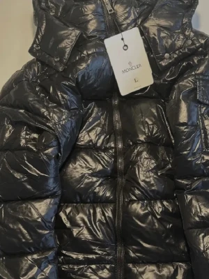 Svart Moncler maya pufferjacka L - Svart pufferjacka från Moncler i storlek L. Jackan har glansig finish, huva och dragkedja framtill. Klassisk Moncler-logga på bröstet och coolt innerfoder med serietidningsmotiv. Perfekt för dig som vill ha en fet vinterjacka med street vibe. Nfc tagen funkar och scanner. Med