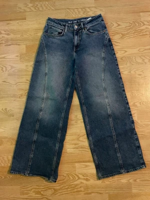 Wide jeans från Bikbok - Säljer ett par blå jeans från Bikbok med breda ben och coola söm-detaljer längs framsidan. Modellen är midwaist och har klassiska fem fickor, bälteshällor och knappstängning. Materialet är jeans och färgen är mörkblå med lätt tvättad look.