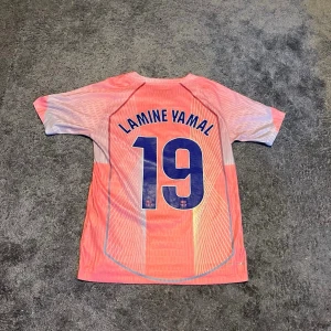 Lamine Yamal #19 Fc Barcelona - Bilderna visar en fotbollströja från FC Barcelona med Lamine Yamal och nummer 19 på ryggen, tillverkad av Nike. Tröjan har ett mönster i rosa och ljusblå toner samt sponsorlogga från Spotify på framsidan.