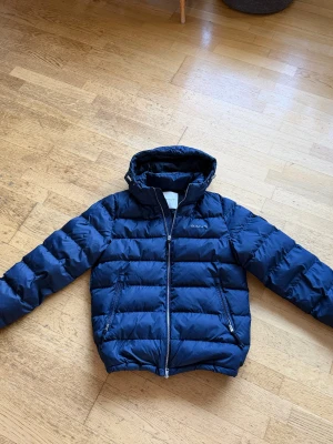 Mörkblå pufferjacka från GANT - Snygg mörkblå pufferjacka från GANT med huva och dragkedja framtill. Jackan har två sidofickor med dragkedja och broderad logga på bröstet. Den är quiltad och har en loose passform, perfekt för kalla dagar. Nypris 3500:-, pris går att diskutera vid snabb affär