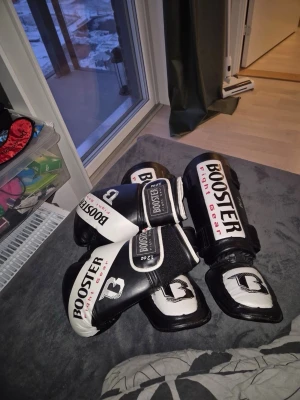 Booster Fight Gear boxningshandskar & benskydd - Säljer ett set med svarta och vita boxningshandskar (12 oz) och benskydd från Booster Fight Gear. Handskarna har bred kardborrestängning och är tillverkade i syntetmaterial. Benskydden har justerbara remmar och förstärkt vaddering för extra skydd vid träning.