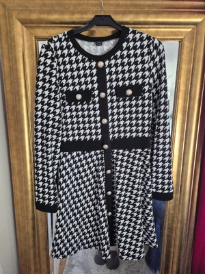 Svartvit rutig kortklänning med knappar - Snygg svartvit kortklänning med klassiskt hundtandsmönster, dekorativa guldfärgade knappar framtill och på fickorna. Klänningen har rund hals, långa ärmar och markerad midja med svarta detaljer. Perfekt för dig som gillar retrostil och vill sticka ut.
