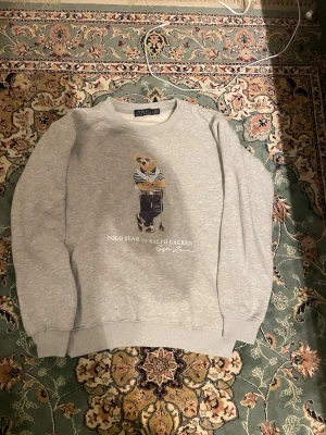 Grå sweatshirt Polo Bear Ralph Lauren - Säljer en grå sweatshirt från Ralph Lauren med Polo Bear-tryck på bröstet. Tröjan har rund hals, ribbade muddar och är långärmad. Materialet är mjuk bomull och passformen är normal. Perfekt för chill dagar och snygg streetstyle.