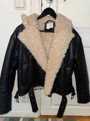 Svart teddyjacka från H&M med bälte - Säljer en svart teddyjacka från H&M i storlek XS. Jackan har en fluffig beige teddykrage och foder, samt detaljer som bälte och dragkedjor vid ärmsluten. Yttermaterialet är syntet med skinnlook och jackan har en loose passform.