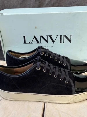 Lanvin svarta sneakers med lackad tå - Säljer ett par stilrena svarta sneakers från Lanvin med snygg lackad tå och ovandel i mocka. Skorna har snörning och vit platt sula som ger en clean look. Perfekta för dig som gillar exklusiva detaljer och fransk design.