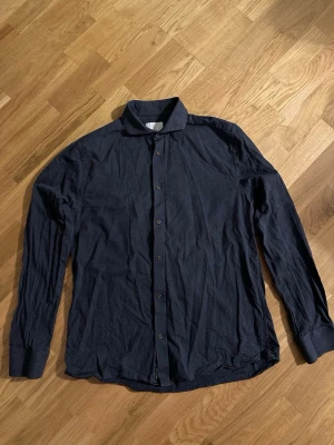 Mörkblå skjorta från Bläck - Snygg mörkblå skjorta med klassisk krage och knappar hela vägen framtill. Skjortan har långa ärmar med breda muddar och är gjord i ett mjukt bomullstyg. Perfekt för dig som gillar enkel och stilren design.