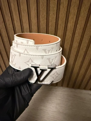 Bälte  - Säljer ett snyggt vitt bälte med klassiskt monogrammönster. Bältet är tillverkat i präglat skinn och har en exklusiv känsla. Perfekt för att lyfta din outfit med en lyxig touch.